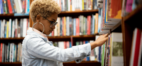 young twenty something female student in denim jacket looking through books on a shelf in a library